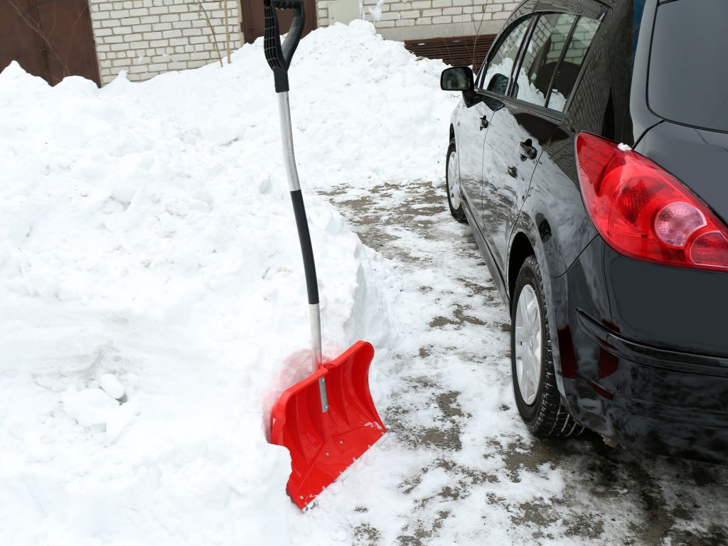 Red snow shovel beside a car on a snow-covered concrete driveway during winter.