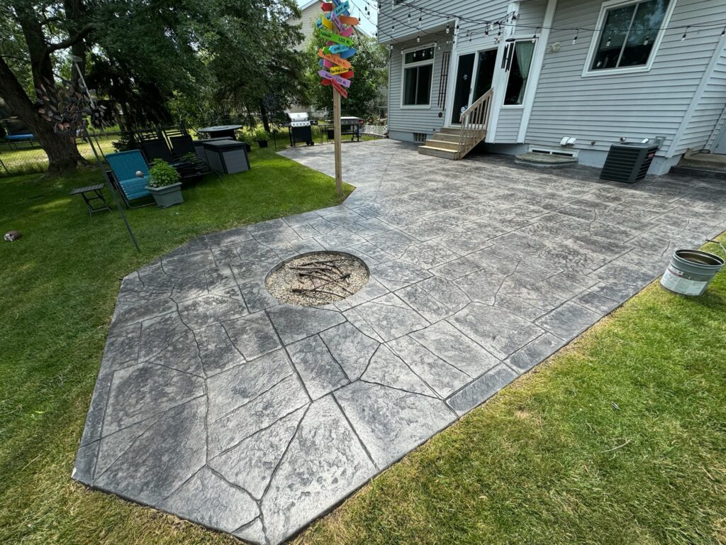 Decorative stamped concrete patio with a stone pattern and built-in fire pit, adjacent to a house and backyard.